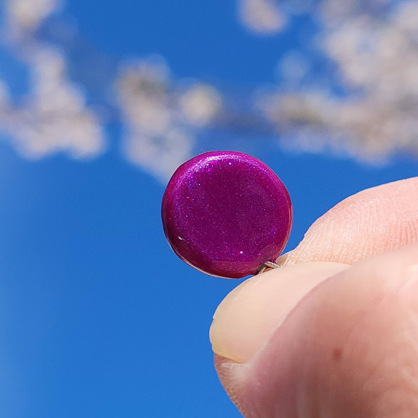 This image showcases a handmade NFC charm being held delicately between two fingers against a bright blue sky. The charm is a deep, sparkling purple with a high-gloss, reflective finish that catches the sunlight beautifully, creating a rich, jewel-toned shine. The background is softly blurred with hints of cherry blossoms or spring foliage, emphasizing the charm’s vibrant color and small size.